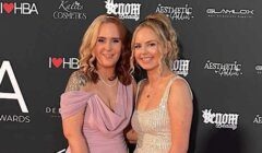 Two women smiling and posing together at an event, standing in front of a step and repeat backdrop displaying various brand logos.