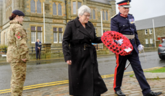 A woman and a man in military uniform hold a wreath of red poppies during a ceremony, with a soldier and a stone building in the background.