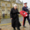 A woman and a man in military uniform hold a wreath of red poppies during a ceremony, with a soldier and a stone building in the background.