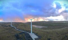 A landscape featuring multiple wind turbines on a hilly terrain during sunset, with a winding road and a small body of water in the foreground.