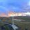 A landscape featuring multiple wind turbines on a hilly terrain during sunset, with a winding road and a small body of water in the foreground.