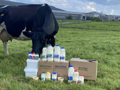 A cow stands in a grassy field next to various milk and dairy product containers, including milk bottles, cream tubs, and a box labeled "Butter." A farm is visible in the background.