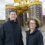 Two people in black jackets stand outdoors in front of industrial equipment with height markings.