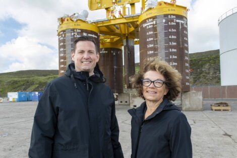 Two people in black jackets stand outdoors in front of industrial equipment with height markings.