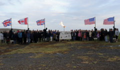 A large group of people stands outdoors by a sign reading "DDAY80", with three British flags, one Canadian flag, and two American flags displayed, and a torch-lit at the center.