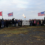 A large group of people stands outdoors by a sign reading "DDAY80", with three British flags, one Canadian flag, and two American flags displayed, and a torch-lit at the center.