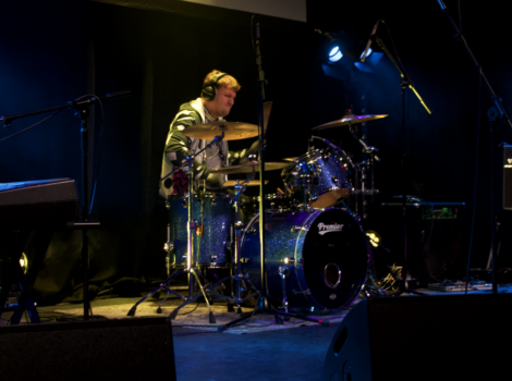 A drummer wearing headphones plays a drum kit on a dimly lit stage, with blue spotlights illuminating the background.