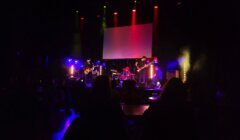 A live band performs on a dimly lit stage with colorful lighting. An audience watches from the shadowy seating area in the foreground. A large screen is positioned behind the band.