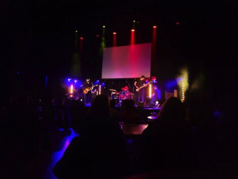 A live band performs on a dimly lit stage with colorful lighting. An audience watches from the shadowy seating area in the foreground. A large screen is positioned behind the band.