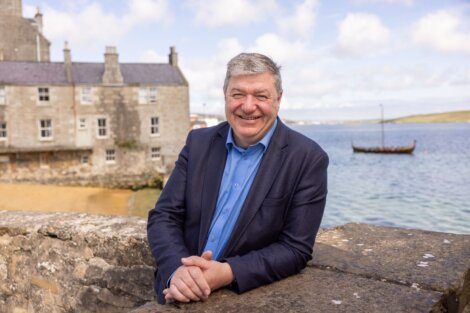 A man in a blue shirt and dark jacket stands smiling with folded hands on a stone wall. Behind him is a waterfront scene with stone buildings, calm water, and a small boat in the distance.