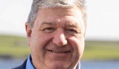 A man with short gray hair is smiling while wearing a blue shirt and a blazer. The background shows an out-of-focus natural landscape.