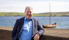 A man in a blue shirt and dark blazer leans against a stone wall by the water, with a Viking longship in the background on a sunny day.