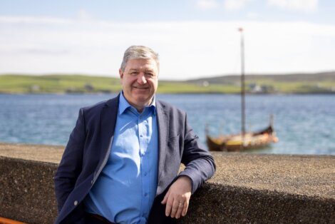 A man in a blue shirt and dark blazer leans against a stone wall by the water, with a Viking longship in the background on a sunny day.