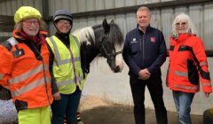 Four people in high-visibility clothing stand in a stable next to a black and white horse.