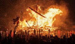 A large wooden structure, resembling a Viking ship, is engulfed in bright flames while an audience watches in the dark, holding torches and capturing the scene with phones.