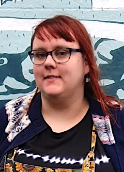 A person with red hair and glasses is standing in front of a mural wearing a dark top and patterned outerwear.