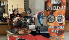 Two children in Viking costumes stand behind a display table with shields, an axe, a dragon head, and a laptop. A banner beside them reads "Junior Up Helly Aa - Find Out More Here.