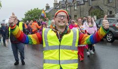 A person wearing a yellow safety vest and rainbow clothing raises both thumbs up while leading a group of people participating in a cheerful parade on a cloudy day.