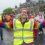 A person wearing a yellow safety vest and rainbow clothing raises both thumbs up while leading a group of people participating in a cheerful parade on a cloudy day.