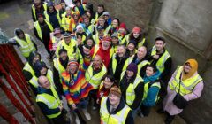 A large group of people in high-visibility vests gather outside a building, looking up at the camera and smiling. Some are dressed in colorful clothing and rain gear.
