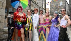 A group of eight people in colorful, flamboyant costumes stand together on a narrow street with stone buildings. One person holds a rainbow umbrella. Bunting is visible in the background.