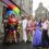 A group of eight people in colorful, flamboyant costumes stand together on a narrow street with stone buildings. One person holds a rainbow umbrella. Bunting is visible in the background.