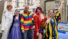 A group of people in colorful and festive outfits pose together on a street. Some wear bright costumes and wigs, and one person is dressed in a rainbow suit. Flags and buildings are in the background.