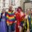 A group of people in colorful and festive outfits pose together on a street. Some wear bright costumes and wigs, and one person is dressed in a rainbow suit. Flags and buildings are in the background.