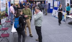 People attending an indoor expo, engaging in conversations with various informational posters and display booths in the background.