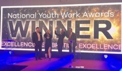 Four people celebrating on stage at the National Youth Work Awards event, standing before a large banner reading "WINNER EXCELLENCE CELEBRATION EXCELLENCE".