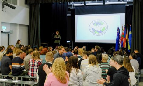 A presenter speaks at a podium on a stage with a screen displaying "The Global Classroom Partnership" logo. An audience sits and some members clap. Flags are positioned on the right side of the stage.