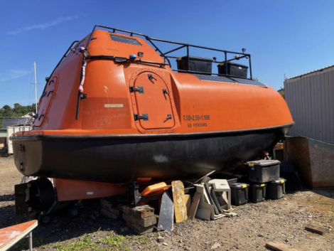 An orange enclosed lifeboat is placed on wooden blocks and storage bins outdoors. It has multiple latches and windows, with part of a building visible on the right and a sailboat mast in the background.