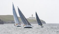 Three sailboats navigate choppy waters near a lighthouse situated on a grassy cliff.