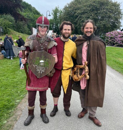 Three people dressed in medieval-style costumes pose together outdoors. One holds a shield, another wears a yellow and red outfit, and the third holds a lyre. They stand on a paved path with greenery behind them.