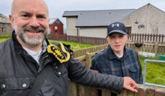 Two men standing outdoors next to a wooden fence with small houses in the background. One man is smiling and carrying a bag, while the other is wearing a hat and leaning on the fence.