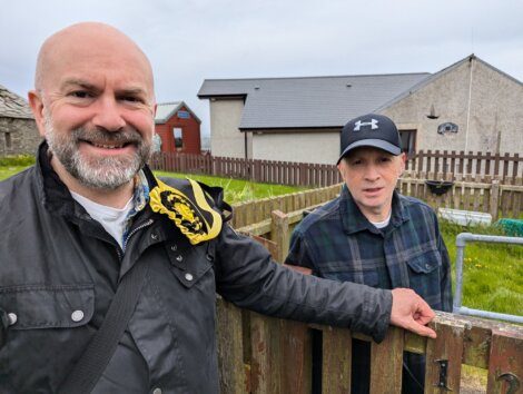 Two men standing outdoors next to a wooden fence with small houses in the background. One man is smiling and carrying a bag, while the other is wearing a hat and leaning on the fence.