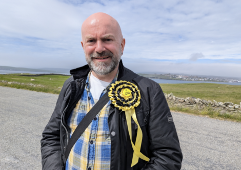 A man with a bald head and beard stands outdoors. He is wearing a black jacket over a blue and yellow plaid shirt and has a yellow and black rosette pinned to his jacket. The background shows a rural landscape.