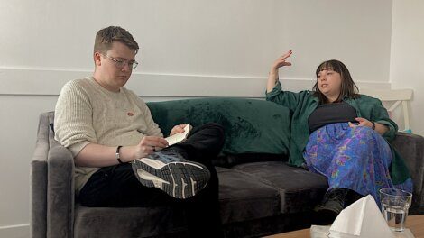 Two people are seated on a couch in a living room. One person is taking notes in a notebook, while the other person is gesturing with their hand as they talk. A box of tissues is on the table in front.