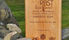 Wooden plaque with the text "RBST Scotland Food & Farming Sustainability Awards 2024, Sustainable Farm of the Year, Uradale Farm" next to a curved horn, set against a rural landscape background.