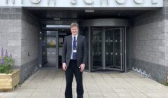 A person stands smiling outside the entrance of a high school building.