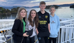 Four teenagers standing on a boat deck with water and islands in the background during sunset. They are all looking at the camera, with two boys on the right and two girls on the left.