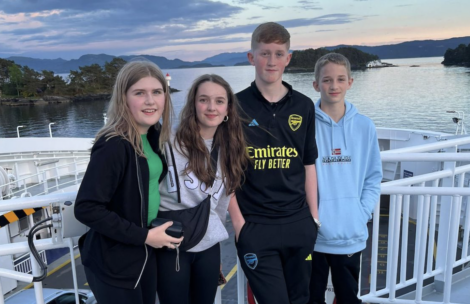 Four teenagers standing on a boat deck with water and islands in the background during sunset. They are all looking at the camera, with two boys on the right and two girls on the left.