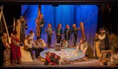 A group of actors in elaborate historical costumes perform a stage play featuring a large, vibrant circular prop at the center, with some standing, sitting, or kneeling around it. Blue backdrop with trees.