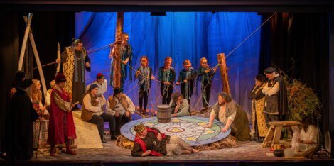 A group of actors in elaborate historical costumes perform a stage play featuring a large, vibrant circular prop at the center, with some standing, sitting, or kneeling around it. Blue backdrop with trees.