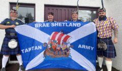 Four men in kilts hold a "Brae Shetland Tartan Army" banner with Viking characters in front of two maroon doors.