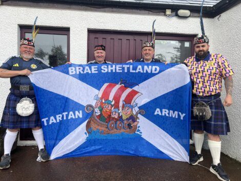 Four men in kilts hold a "Brae Shetland Tartan Army" banner with Viking characters in front of two maroon doors.