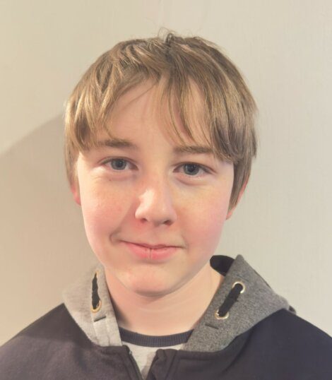 A young person with short, light-brown hair wears a grey and black jacket. They look directly at the camera, smiling slightly, standing against a plain beige background.