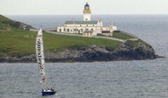 A sailboat with a large white sail is visible in the foreground on the water, with a lighthouse on a rocky hill in the background.