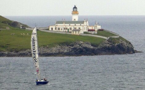 A sailboat with a large white sail is visible in the foreground on the water, with a lighthouse on a rocky hill in the background.