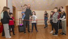 A group of people, including children, are in a room with wooden flooring and disco lights. A man holding a toddler stands next to a girl holding a red ribbon, while others watch and smile.